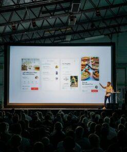 person discussing while standing in front of a large screen in front of people inside dim-lighted room