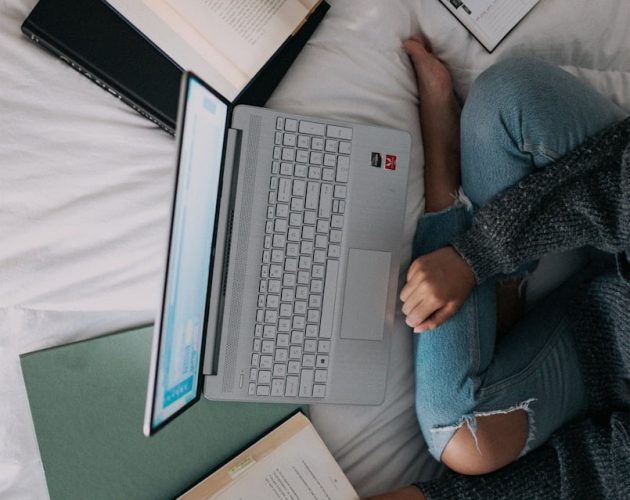 woman in blue long sleeve shirt and blue denim jeans sitting on bed using laptop